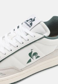 Fehér sneaker zöld részletekkel, texturált felsőrésszel, velúr elemekkel és logóval a nyelvén. A fűző fehér és lapos.