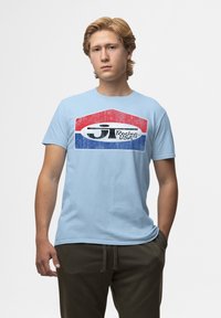 Světle modré bavlněné tričko s vintage grafikou v červené, bílé a modré barvě s textem "Racing USA" a geometrickým designem.