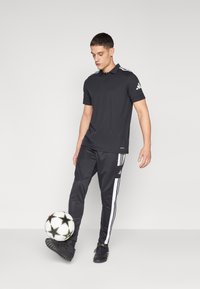 Svart Adidas polo skjorte med korte ermer, kombinert med svarte bukser med hvite sidestriper. En svart fotball holdes nær foten.