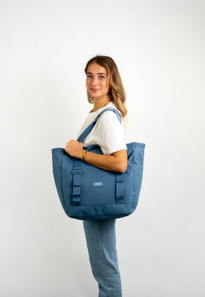 Blaues Tragetasche aus strukturiertem Stoff mit zwei Griffen, einem auffälligen Logo auf der Vorderseite und einer breiten, rechteckigen Form.