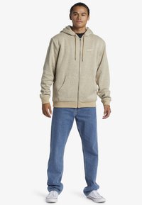 Felpa beige con zip, tasca frontale, cappuccio con coulisse e polsini a costine. Abbinata a jeans azzurri a gamba dritta e sneakers bianche.