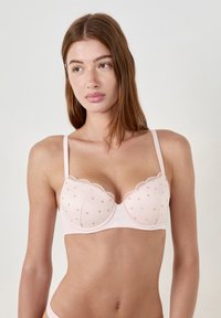 Jasnoróżowy biustonosz z falbaniastymi brzegami, w kropeczki, z fiszbinami i cienkimi regulowanymi ramiączkami. Miękka tekstura materiału.