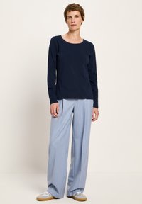 Frau mit kurzen braunen Haaren, die ein marineblaues Langarmoberteil, hellblaue weite Hosen und weiße Turnschuhe trägt, steht vor einem schlichten Hintergrund.