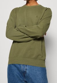 Osoba ze skrzyżowanymi ramionami, ubrana w oliwkowo-zielony, teksturowany sweter z długim rękawem oraz niebieskie dżinsy, na jednolitym tle.