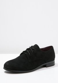 Chaussure oxford en suède noir avec un bout rond, un laçage à l'avant et un petit talon carré. Texture lisse et design minimaliste.