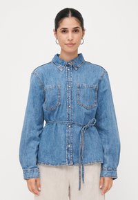 Camisa de ganga azul com botões, de mangas longas, duas bolsos no peito e um cinto correspondente à cintura, apresentando costuras em contraste.