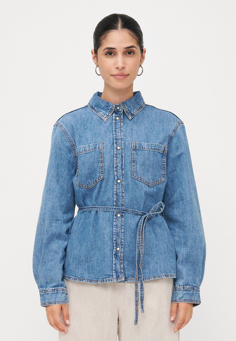 Camisa de ganga azul com botões, de mangas longas, duas bolsos no peito e um cinto correspondente à cintura, apresentando costuras em contraste.