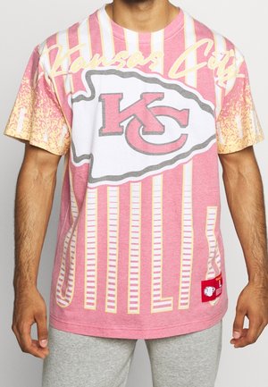 Uomo che indossa una maglietta rosa dei Kansas City Chiefs con un grande logo della squadra e strisce verticali, abbinata a pantaloni grigio chiaro.