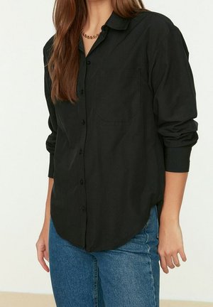 Femme portant une chemise noire boutonnée avec les manches retroussées et un jean bleu, debout devant un fond clair uni.