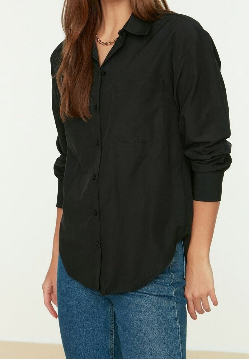 Femme portant une chemise noire boutonnée avec les manches retroussées et un jean bleu, debout devant un fond clair uni.