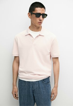 Jeune homme portant des lunettes de soleil noires rectangulaires, un polo rose pâle à manches courtes sur un t-shirt blanc, et un jean bleu, debout devant un fond uni.