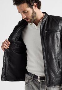 Sort læderjakke med lynlåse foran, stående krave og lynlåslomme på brystet, båret åben over en hvid polo-shirt. Indersiden er sort.