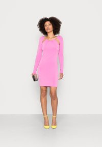 Vestido rosa de mangas longas com detalhes recortados, design justo e textura suave. Combinado com sapatos amarelos de bico fino e uma carteira.