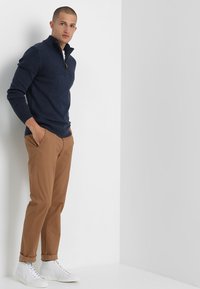 Tummansininen neulepusero, jossa on puolivetoketjukaulus, khakin väriset slim-fit-housut ja valkoiset lenkkarit. Sileä rakenne ja rento muotoilu.