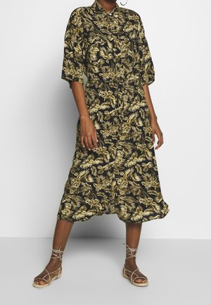 Mujer con un vestido midi negro con estampados de hojas tropicales beige y sandalias planas beige con tiras, de pie contra un fondo liso.