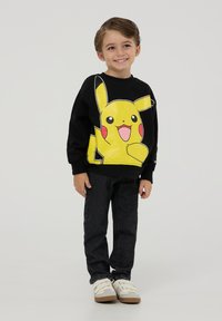 Czarny sweatshirt z dużą żółtą grafiką Pikachu, noszony z czarnymi dżinsami i białymi butami, stojący na jednolitym tle.
