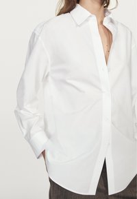 Camicia bianca con bottoni realizzata in tessuto fresco, con colletto classico, maniche lunghe e una vestibilità rilassata con orlo curvo.
