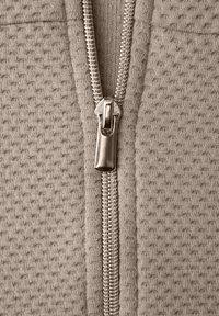 Detailní záběr na béžovou texturovanou látku se stříbrným kovovým zipem uprostřed, který je částečně rozepnutý.