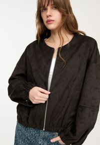 Svart mockabomberjacka med rund halsringning, dragkedja fram, elastiska cuffs och en rynkad nederkant. Bärs över mönstrad denim.