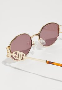 Gafas de sol redondas con lentes rosa, marco de metal dorado y varillas adornadas con un logo circular. Almohadillas nasales transparentes y detalles de carey en las patillas.