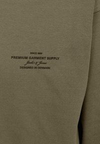 Olíva zöld pulóver, amely puha tapintású, fekete nyomtatott szöveggel. Részletek között szerepel a "Premium Garment Supply" és a "Designed in Denmark."