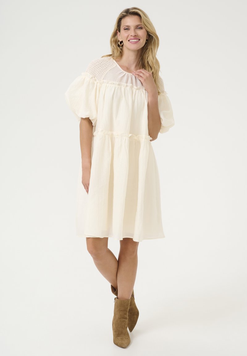 Eine Frau lächelt und trägt ein lockeres, cremefarbenes Kleid mit Puffärmeln und braunen Wildleder-Ankle-Boots, während sie vor einem schlichten weißen Hintergrund steht.