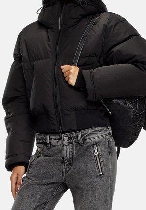 Osoba w czarnej kurtce puchowej i szarych dżinsach z kieszeniami na zamek, niosąca czarny plecak na jednym ramieniu.