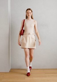 Mini-jupe beige avec poches latérales, débardeur ajusté et sac à main rouge. Le modèle porte des chaussettes blanches et des chaussures rouges, se tenant devant un mur clair.