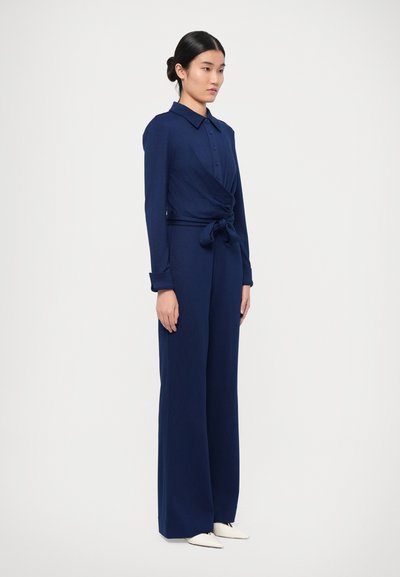 Marineblauwe jumpsuit met een knoopsluiting bij de kraag, lange mouwen en een geknoopt tailleband. Kenmerkt zich door wijde benen en een gladde stofstructuur.