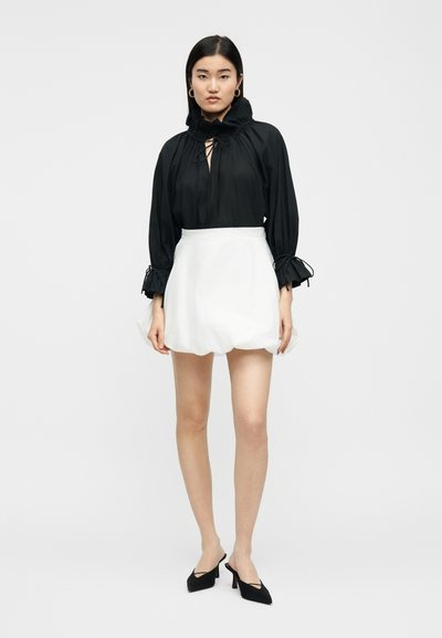 Γυναίκα που φοράει μαύρη μπλούζα με μακριά μανίκια, βολάν στα μανίκια και το γιακά, λευκή μίνι φούστα με φουσκωτή τελειώματα και μαύρα μυτερά τακούνια.