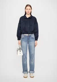 Marineblå kort jakke med glidelås, elastisk kant; lyseblå jeans med X-mønster, rullet ermer; hvit veske med svarte detaljer; beige sneakers.