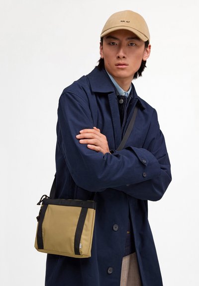 Jeune homme portant une casquette beige, un manteau bleu marine et un sac bandoulière marron clair, debout les bras croisés devant un fond blanc uni.