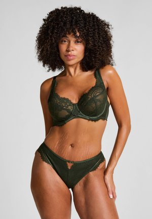 Mujer con cabello rizado que lleva un sujetador de encaje verde y bragas a juego, de pie contra un fondo gris claro liso.