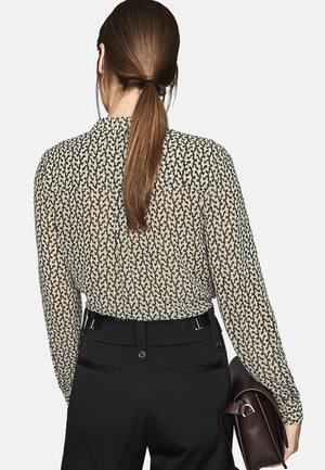 Femme avec une queue de cheval brune portant un chemisier à manches longues à motifs rentré dans un pantalon noir taille haute, tenant un sac à main en cuir marron foncé.