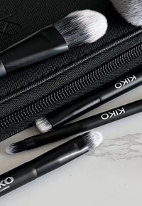 Set med KIKO sminkborstar med svarta skaft och mjuka, vita borsttoppar, arrangerade på ett texturerat svart fodral med dragkedja, som framhäver deras design.