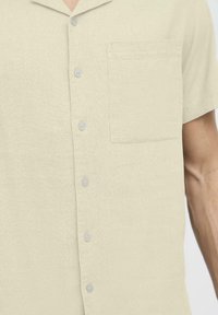 Chemise courte à manches courtes de couleur beige clair en tissu texturé, dotée d'une fermeture à boutons, d'une poche poitrine et de boutons gris.