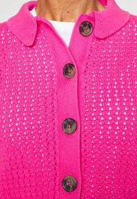 Cardigan en tricot rose vif avec de gros boutons foncés boutonnés sur une chemise blanche, montrant un col.