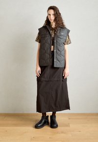 Gilet trapuntato nero con bottoni a scatto, abbinato a una blusa a maniche corte con fantasia e una lunga gonna nera, indossato con stivaletti neri alla caviglia.
