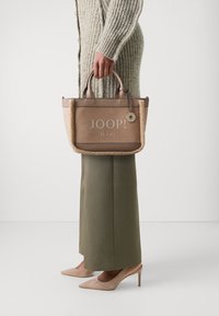 Torebka w odcieniach brązowym i beżowym z teksturowaną powierzchnią, z wyraźnym logo "JOOP! JEANS". Krótkie uchwyty i ozdobny charms jako akcent.