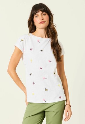 Frau mit langen braunen Haaren, die ein weißes T-Shirt mit kleinen bunten Palmenmotiven und grüne Hosen trägt, steht mit einer Hand auf der Hüfte.