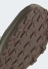 Gummisohle mit ausgeprägten Profilstollen, einer strukturierten Oberfläche und hexagonalem Muster. Die Farben umfassen Braun-, Grau- und Olivtöne.