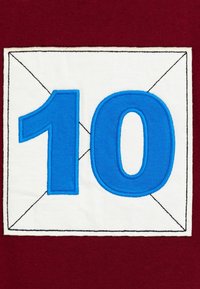 Kék 10-es szám hímezve egy fehér négyzet alakú folton, fekete varrású kerettel, sötétvörös szövet háttéren.
