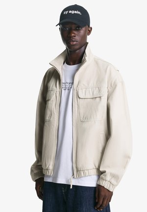 Young man wearing a beige zip-up jacket, white printed t-shirt, dark jeans, and black cap with "try again." text on a white background.