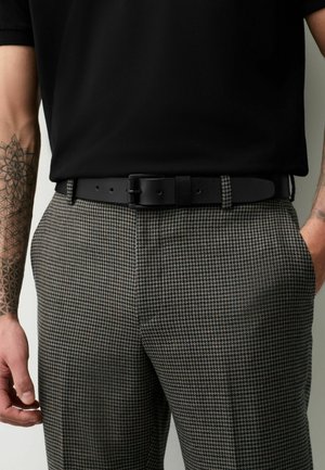 Férfi fekete inget, fekete bőrszíjat és kutyanyakmintás nadrágot visel, tetovált karokat és kezeket mutat a zsebek közelében.