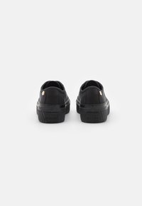 Para czarnych niskich sneakersów z grubymi podeszwami, siateczkowej tkaniny i małymi metalowymi znakami na pięcie, na białym tle.