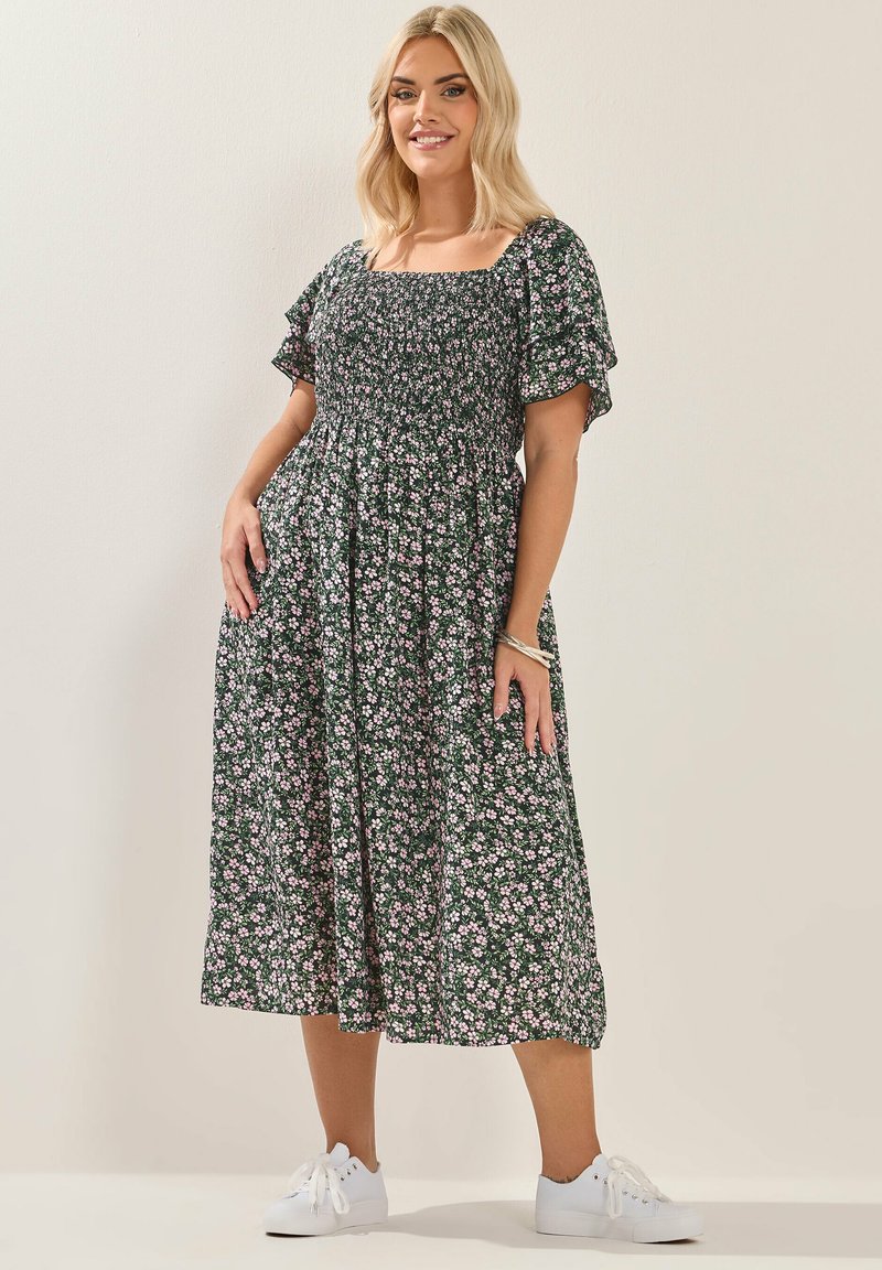 Vrouw met blond haar die een groen bloemenmidi-jurkje en witte sneakers draagt, staand en glimlachend tegen een effen beige achtergrond.