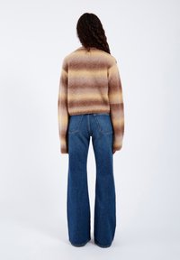 Person med langt krøllet hår iført en brun og beige stribet fluffy sweater og blå jeans med vide ben, stående mod en hvid baggrund.