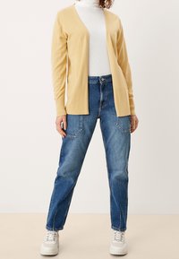 Żółty kardigan z dekoltem w serek i ściągaczami na rękawach, noszony na białym golfie, w połączeniu z luźnymi niebieskimi dżinsami i białymi trampkami.