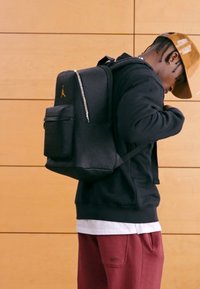 Svart ryggsekk med teksturert overflate, glidelåslomme foran og gyllent logo; båret på ryggen av en person i svart hettegenser.