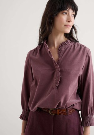 Langsleeve blouse van gedempt mauve stof, met een gerimpelde kraag, knoopsluiting aan de voorkant en elastische manchetten. Gepaard met een gevlochten bruine riem.